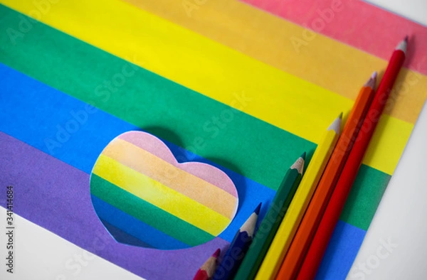 Fototapeta Colored pencils lie on colored paper in the form of a rainbow in the color of the LGBT flag next to a paper colored heart