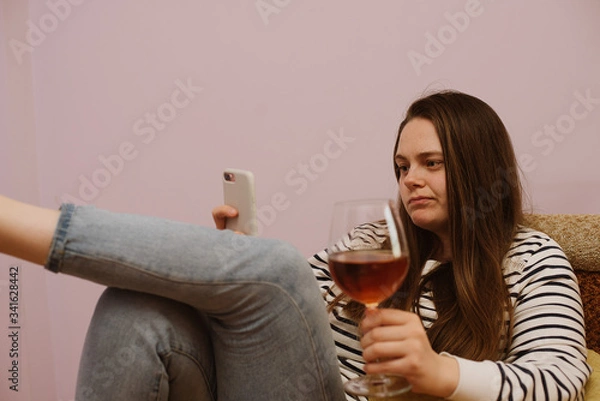 Fototapeta Sad young woman is using phone with glass of wine at home on sofa indoor during home isolation quarantine
