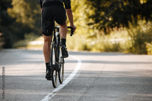 Fototapeta Back view of cyclist in activewear riding professional bike on paved road with blur background of green plants. Concept of active lifestyle and hard training