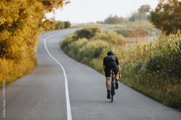 Obraz Widok z tyłu silnego rowerzysty o atletycznej sylwetce jeżdżącego na rowerze przy utwardzonej drodze wśród drzew i zielonych krzewów w czarnym kasku ochronnym i sportowej odzieży. Koncepcja treningu na świeżym powietrzu