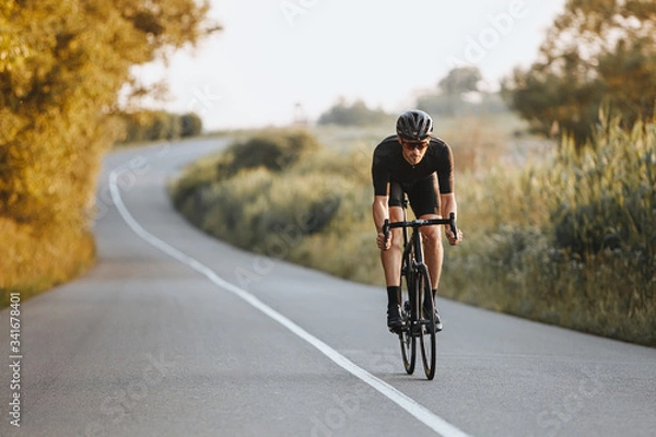 Fototapeta Professional male cyclist in black helmet, protective glasses and activewear dynamically riding bicycle on paved road with blur background. Concept of summer activity and healthy lifestyle