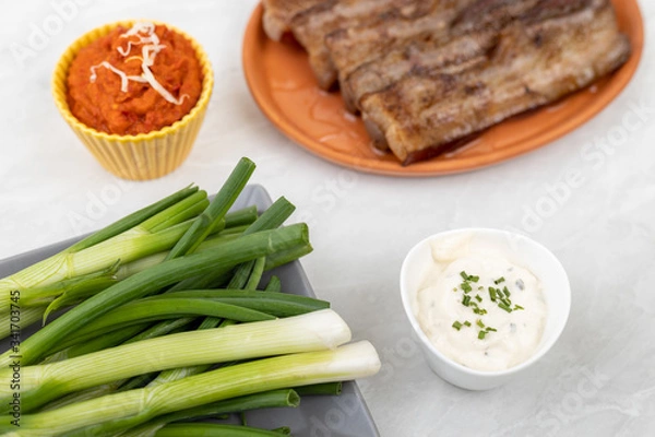 Obraz Young onions on the table with ajvar and tartar sauce with grilled bacon in the background