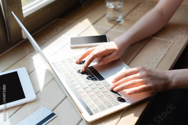 Fototapeta cropped view of woman typing on laptop keyboard near credit card and gadgets with blank screen