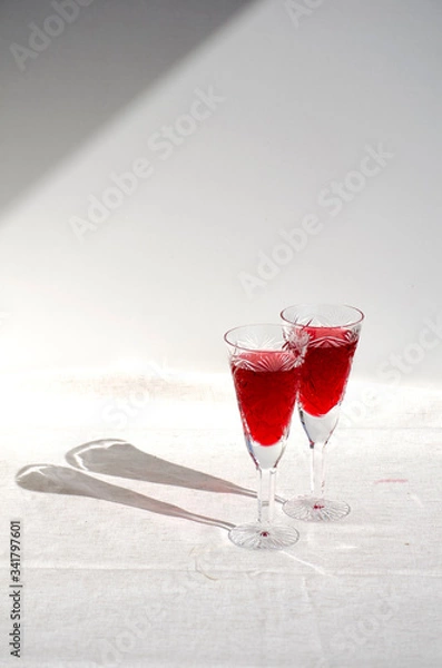Fototapeta Two crystal glasses with red wine. Glass goblets with alcohol on background of wooden wall. White linen tablecloth. Direct sunlight, long shadows. Selective focus image. Copy space.