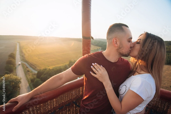 Obraz Beautiful attractive smiling couple staying and kissing in air baloon basket at sunset. Couple first time on baloon flight