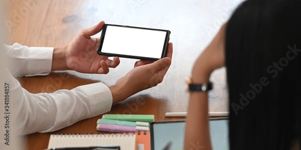 Fototapeta Photo of young businessman showing a white blank screen smartphone in his hands to colleague while sitting together at the wooden working table over orderly workspace as background.