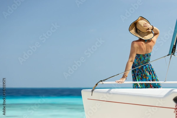Fototapeta Back view of joyful attractive amazing woman in blue dress and hat enjoying azure water on the deck of a sailing yacht. Concept of sailing regatta, luxury vacation at sea.