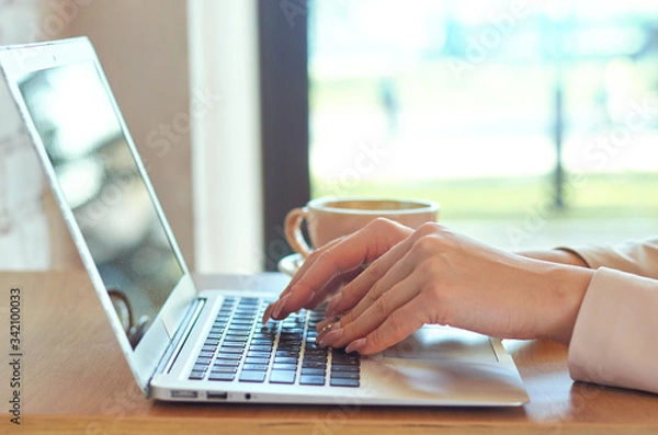 Fototapeta Female young hands with pale pink manicure are typing on a laptop keyboard. Nearby is a fresh mug of coffee.