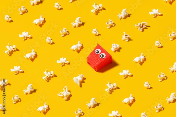 Fototapeta Mini red popcorn box carton with googly eyes on a yellow background surrounded by popped popcorn.