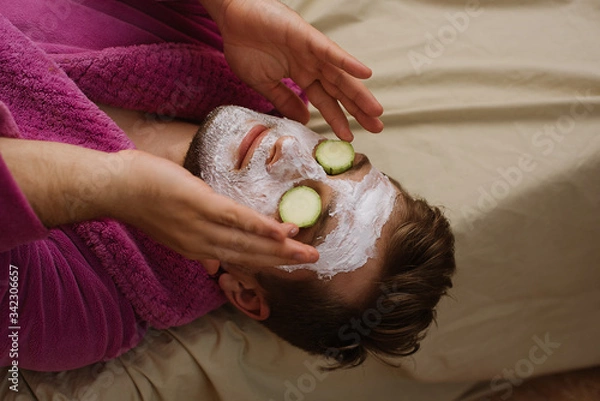 Fototapeta Close up photo of relax man wearing pink robe with facial mask and cucumber on his face laying on the sofa at home isolation quarantine. spa at home concept