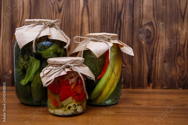 Obraz Homemade pickles in glass jars, cucumbers, peppers. Canned vegetables on a wooden background. Preserves.
