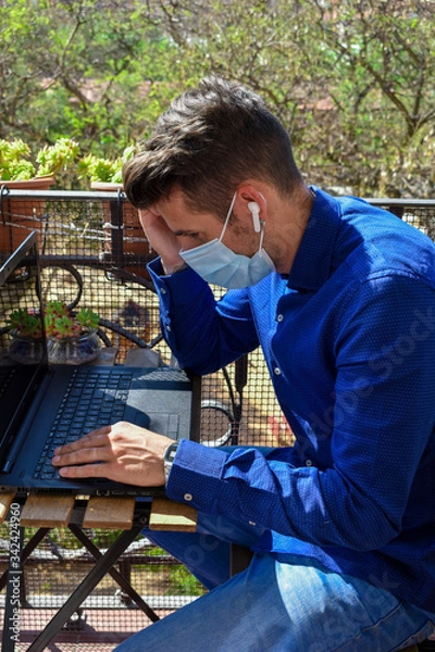 Fototapeta Man wearing a face mask making a video call with the laptop on his balcony, confined at home by the quarantine due to covid-19