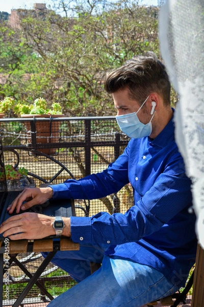 Fototapeta Man wearing a face mask making a video call with the laptop on his balcony, confined at home by the quarantine due to covid-19