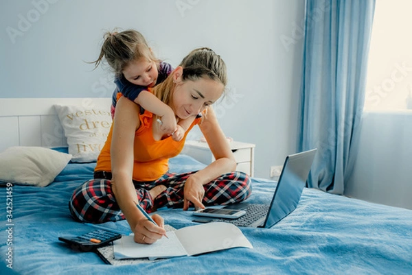 Fototapeta Stay at home mom working remotely on laptop while taking care of her baby. Woman in orange shirt is working office work remotely from home on bed. Distance learning online education and work. 