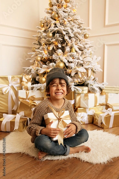 Obraz cute little boy with dark hair in cozy clothes posing near decorated Christmas tree with presents