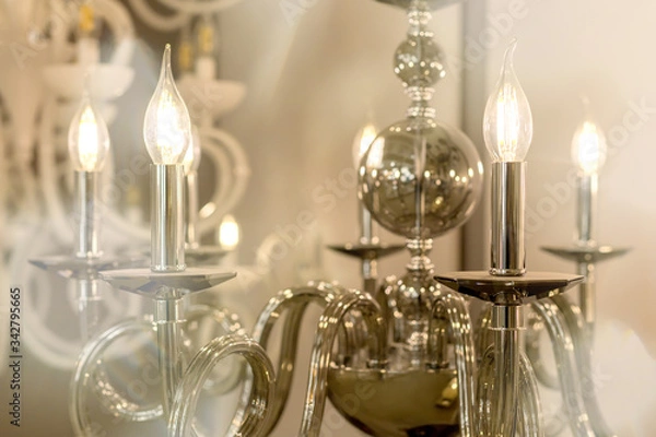 Fototapeta The picture of a glowing chandelier with metal chrome fixtures and shades in the form of candles. The concept of the classical style in the interior. Close up. Soft warm filter, selective focus.