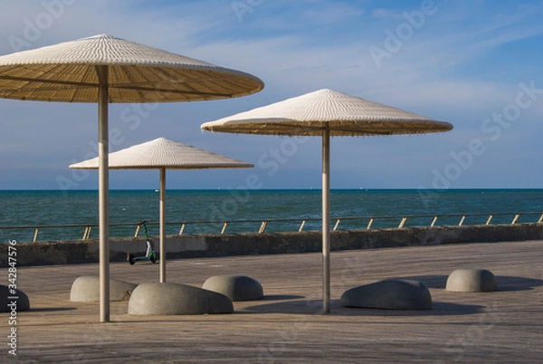 Fototapeta Empty embankment in Tel Aviv, Israel. View of umbrellas and stone benches to relax in a sunny day. The sea and the scooter in the background.