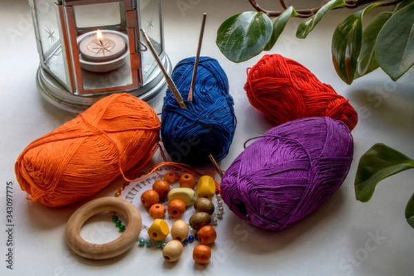 Fototapeta Home hobbies. Composition in bright colors of yarn, beads, pins and knitting needles. Nearby is a candle in a metal candlestick and green leaves of a flower. What to do at home.