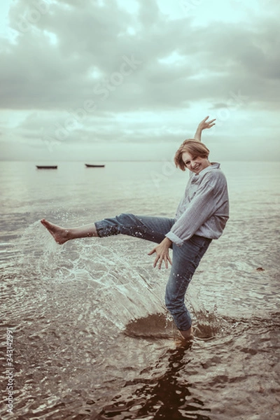 Fototapeta A happy young woman on the lake and expresses her emotions by making a wave with splashes with her leg. A portrait of a smiling female enjoying life. A girl wearing a shirt and blue jeans. Lifestyle