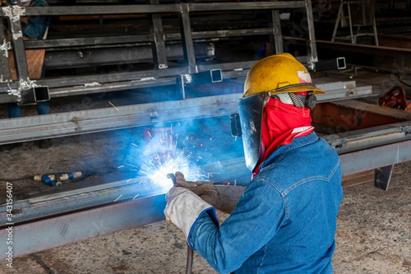 Fototapeta The welder is welding a steel structure work with process Flux Cored Arc Welding(FCAW) and dressed properly with personal protective equipment(PPE) for safety, at industrial factory.