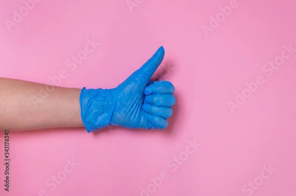 Fototapeta Doctor hand in blue disposable medical glove showing thumbs up on pink background. Gesture of approval or acceptance. Hygiene measures to prevent spread of Covid-19 virus. Coronavirus protection.