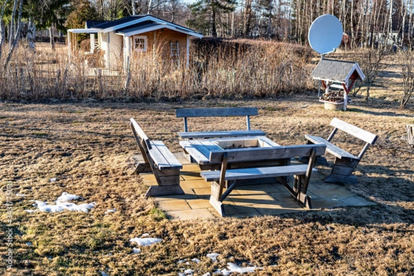 Fototapeta Stylish wooden barbeque spot with benches and table standing at concrete plates in the middle of backyard, spot of snow left at faded after winter grass. Small well and satellite dish at background