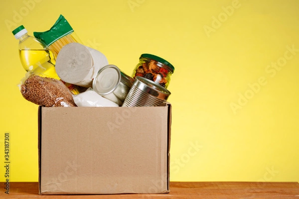 Fototapeta Food Donation concept. Donation Box With food For Donation On Yellow Backround. Assistance To The Elderly In The Context of The Coronavirus Pandemic