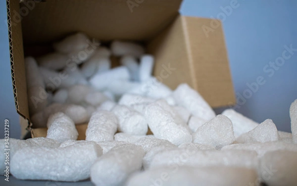 Fototapeta Open cardboard box with white styrofoam filling pieces comming out, parcel packing materials, safe and secure shipment methods, grey background