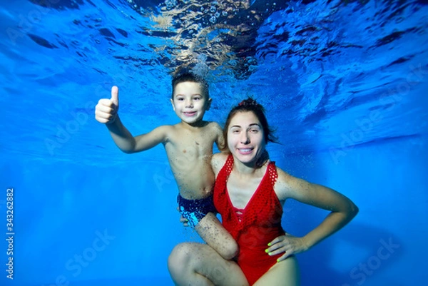Obraz A happy mother and her young son pose for the camera underwater in a pool on a blue background. Mom in a red knitted swimsuit looks at the camera. They smile. Closeup. Digital photo.