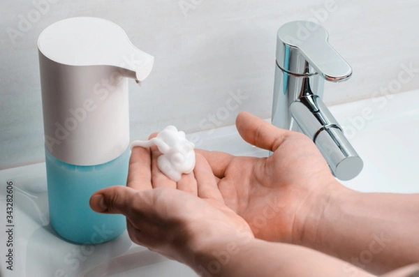 Obraz Automatic soap dispenser in the bathroom. Hands with soapy solution close-up. The fight against coronavirus.