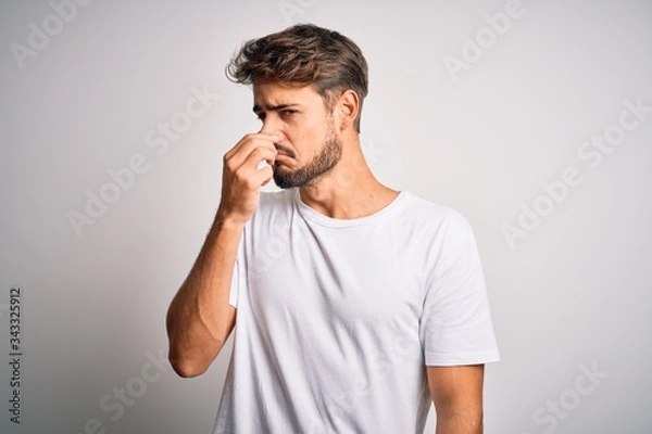 Fototapeta Young handsome man with beard wearing casual t-shirt standing over white background smelling something stinky and disgusting, intolerable smell, holding breath with fingers on nose. Bad smell