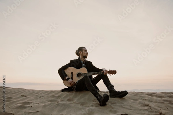 Fototapeta long haired bearded musician with guitar sitting on a sand hill in the sunset