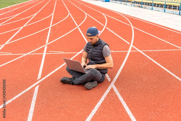 Fototapeta The guy with the laptop sits in the stadium, the programmer in the empty stadium. Working in unusual places