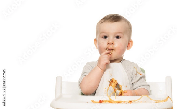 Fototapeta Portrait of funny baby boy that eats spaghetti in tomato sauce on white isolated background.