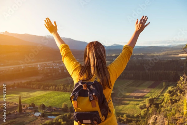 Fototapeta Young woman stands at the top of a hill with her arms raised at golden hour