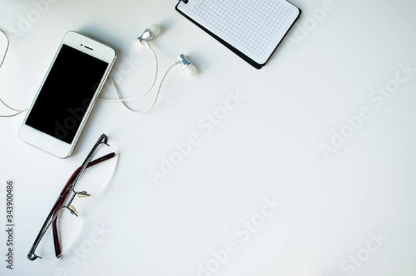 Fototapeta a white phone with headphones and a black screen, glasses and a Notepad. on white background. space for text. Concept of music, recreation, office layout or school and College