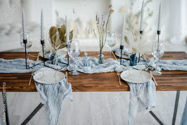Fototapeta Romantic table setting for holiday dinner, wooden table served with dried flower, plates, golden cutlery, white candeles, bright dusty blue runner. Selective focus.