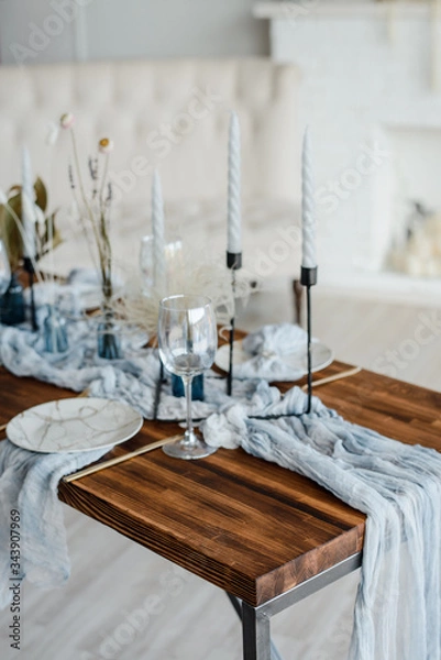Fototapeta Romantic table setting for holiday dinner, wooden table served with dried flower, plates, golden cutlery, white candeles, bright dusty blue runner. Selective focus.