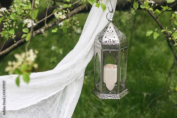 Fototapeta Ornamental silver Moroccan lantern handing on blooming tree. Green blurred background with white veil. Greeting card, invitation for holiday Ramadan Kareem. Garden party wedding deocration