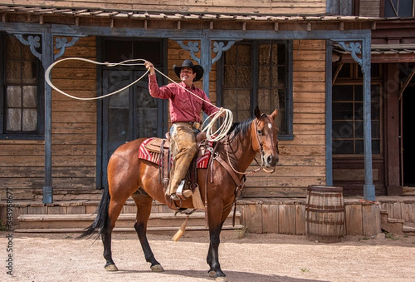 Obraz Cowboy using lasso while riding his horse through an old western town