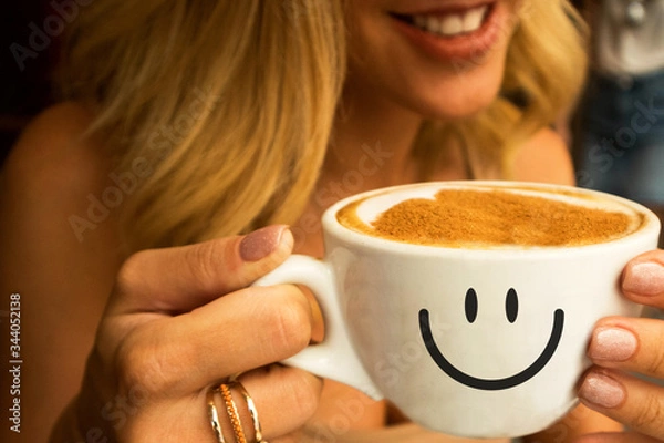 Fototapeta smiling young girl in a cafe  holding a big cup of hot cappuccino with coffee foam design of heart on it and smile on the cup