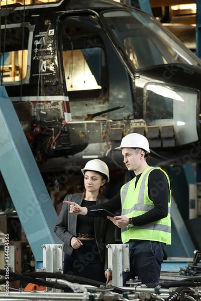 Fototapeta Portrait of a female factory manager in a white hard hat and business suit and factory engineer. Deciding future factory development.  Controlling the work process at the helicopter manufacturer.