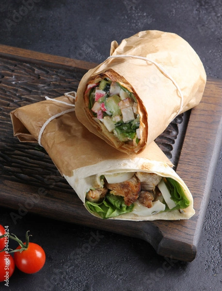 Fototapeta Caesar wrap. Chicken, lettuce, egg, sauce and salad in pita bread on dark wooden board on the black table. Rolls in parchment, cherry tomatoes. Shawarma vertical. Background image, copy space