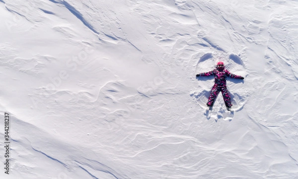 Obraz Top aerial view of young happy smiling girl making by arms snow angel figure and lying in snow, winter outdoor activity concept. Girl in the snow angel shows . Winter angel in a bright ski suit