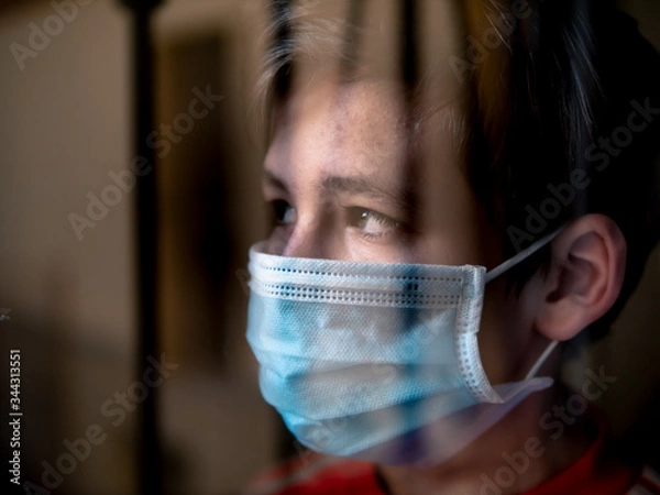 Fototapeta Boy with a medical mask looks sadly out of the window during his confinement because of convid 19