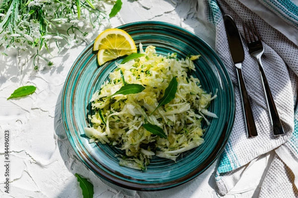 Fototapeta Crispy salad of fresh vegetables with herbs. Young cabbage with cucumber and arugula leaves, decorated with lemon and a bouquet of white flowers on a light textured stone table with Cutlery