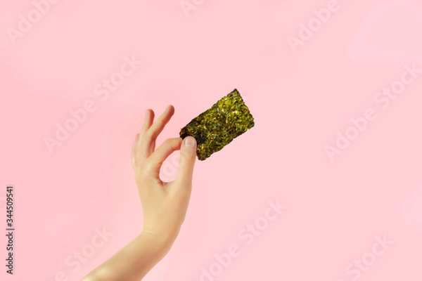 Fototapeta Female hand with crispy seaweed on the pink background with copy space. Dry nori sheets korean or japanese roasted food texture. Flat lay.