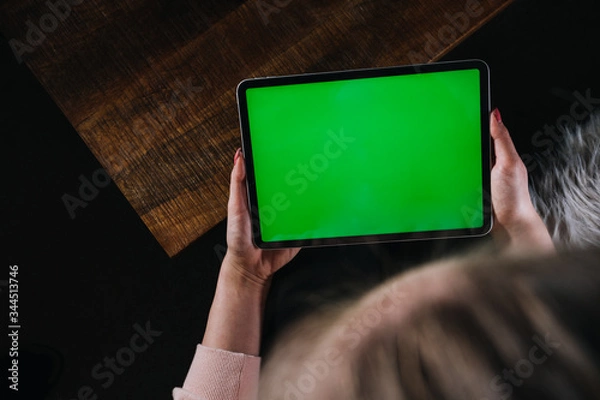 Fototapeta Young woman using black tablet device with green screen. Woman holding tablet, scrolling pages while sitting on the couch in the living room. Chroma key. View over shoulder. Close up