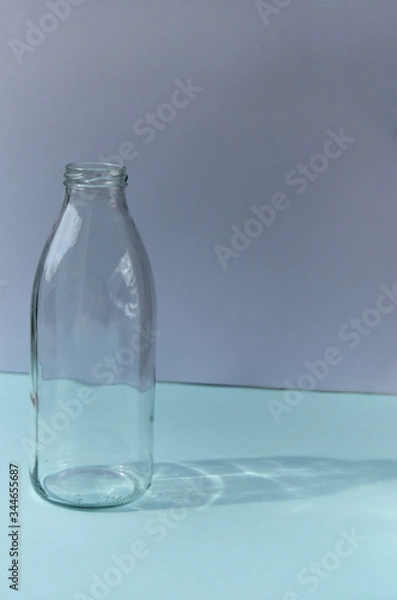 Fototapeta Glass bottle stands on a blue table against the background of a white wall shadow from the bottle sunny