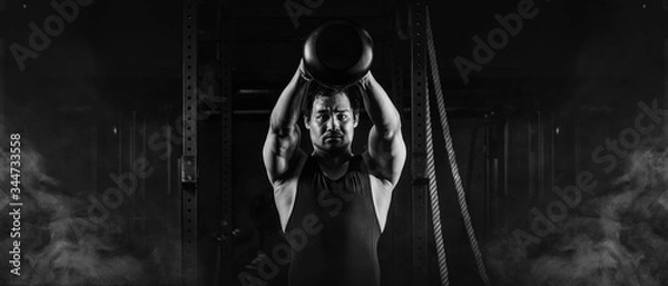 Fototapeta asian athletic strong man having workout and bodybuilding with kettlebell weightlifting backsquat style in gym and fitness center in dark tone black and white